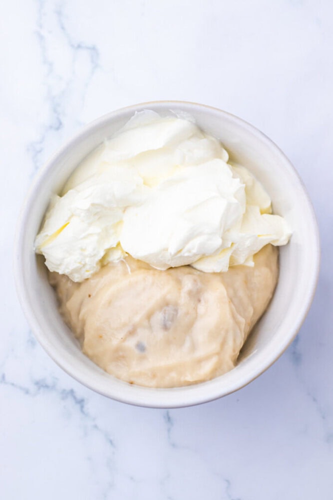 A white bowl containing a mixture of thick, creamy substances, one off-white and the other pale beige, on a light marble surface.