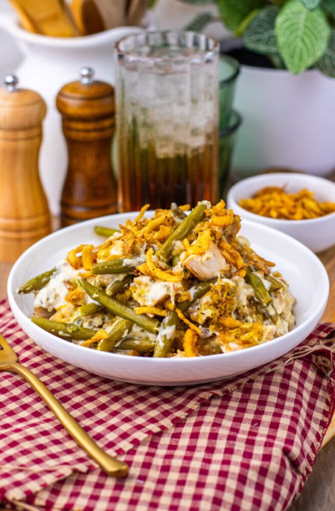 A bowl of green bean casserole topped with crispy fried onions sits on a red checkered cloth, with a drink, pepper mills, and a small bowl of onions in the background.