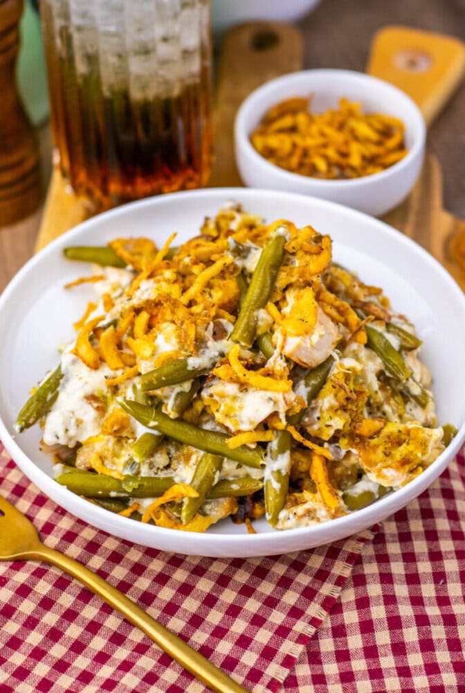 A white bowl filled with green bean casserole topped with crispy fried onions sits on a table with a gold fork, a red checkered napkin, and a small bowl of extra fried onions in the background.