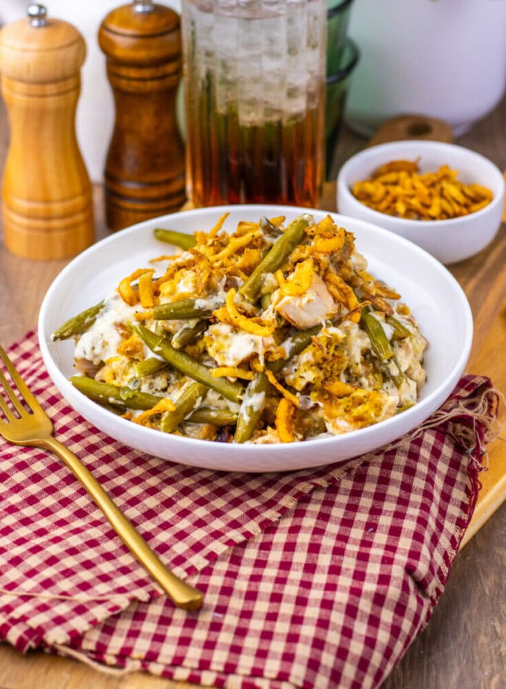 A white bowl filled with a green bean casserole topped with crispy fried onions sits on a red checkered cloth next to a gold fork, with condiments and a drink in the background.