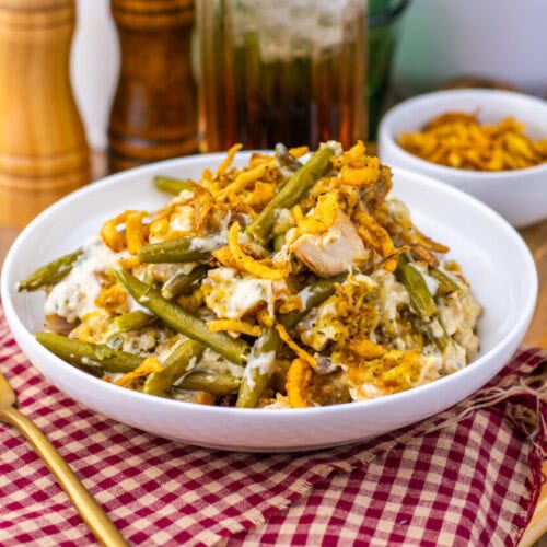 A white bowl of green bean casserole topped with crispy fried onions sits on a red checkered napkin with a fork beside it. Pepper grinders and a drink are in the background.