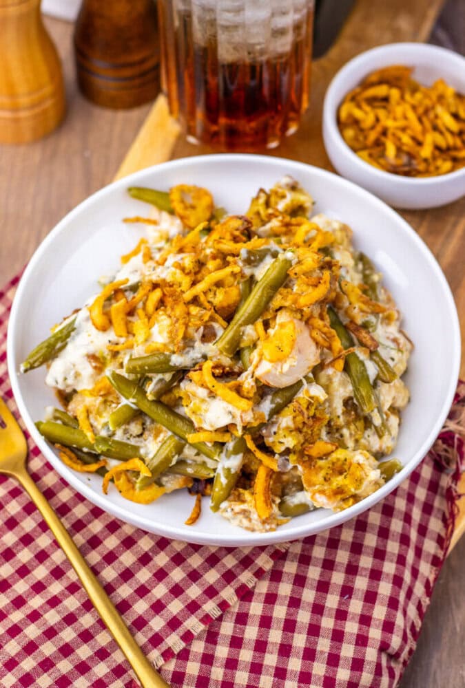 A plate of chicken and green bean casserole topped with crispy onions, set on a red checkered cloth with a fork beside it; a bowl of crispy onions and a drink are in the background.