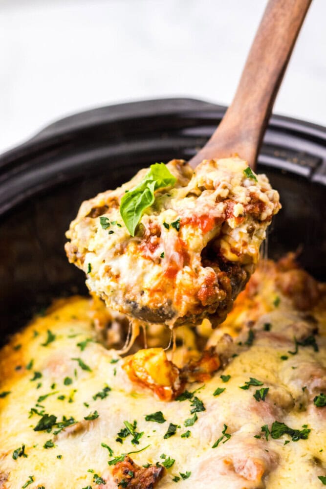 A wooden spoon lifts a cheesy, baked lasagna portion from a slow cooker, topped with herbs and melted cheese.