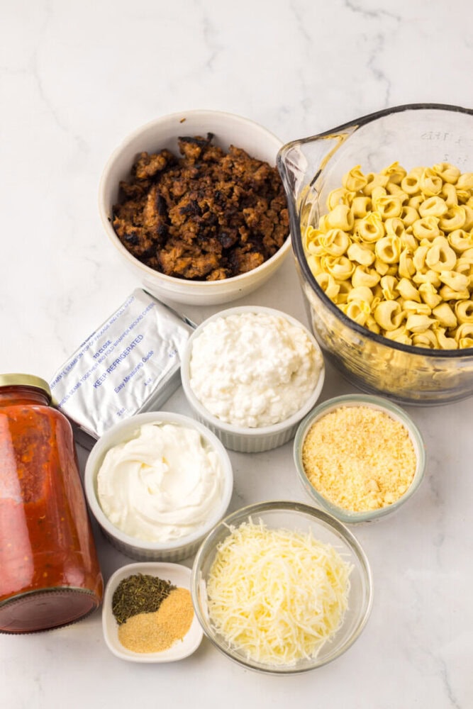 Top-down view of ingredients for a pasta dish, including cooked sausage, tortellini, pasta sauce, cottage cheese, cream cheese, sour cream, shredded cheese, grated cheese, and spices.