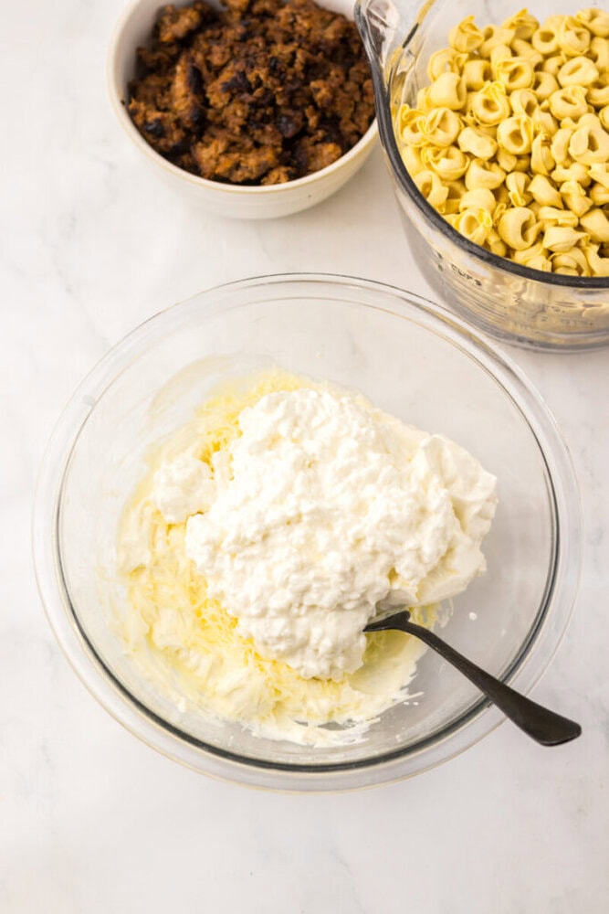 A glass bowl with ricotta cheese mixture and a spoon, next to bowls of cooked tortellini and browned sausage on a white surface.