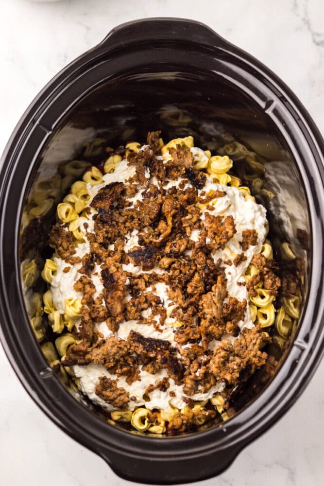 A slow cooker filled with uncooked tortellini, creamy sauce, and browned ground meat on top, ready to be cooked.