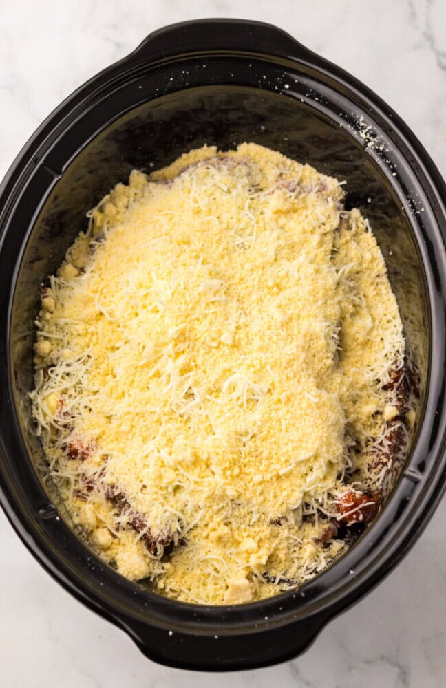 A slow cooker filled with breaded and grated cheese-covered chicken breasts, ready to be cooked.