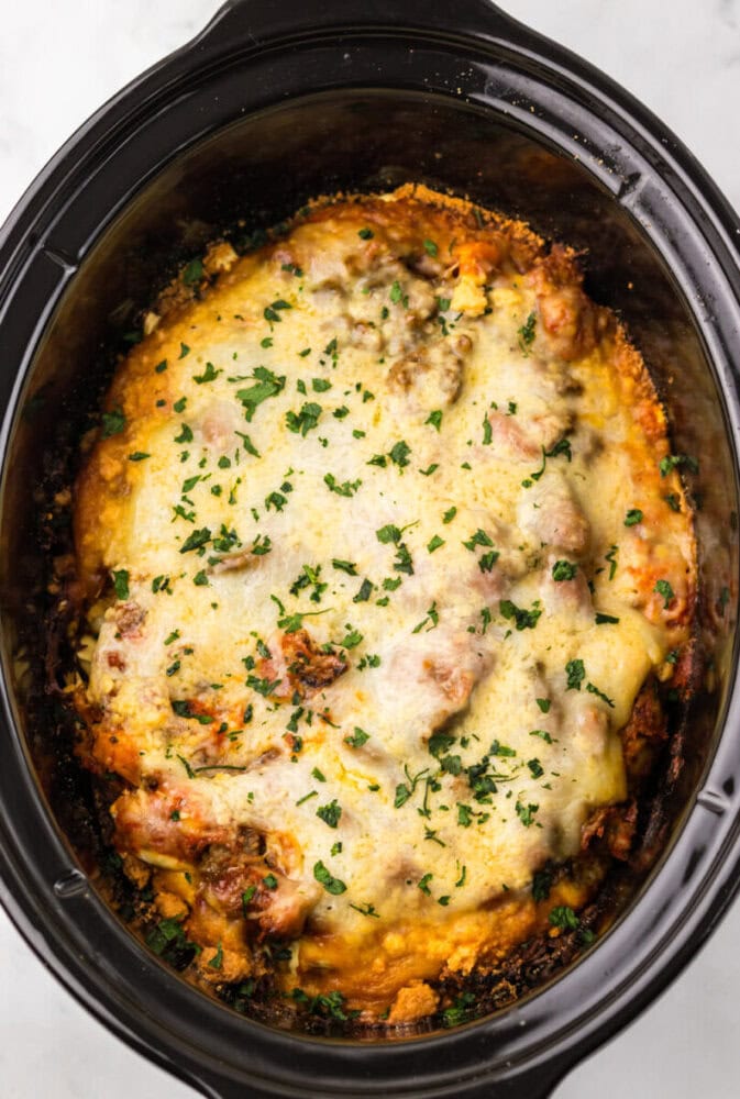 A baked lasagna with melted cheese and chopped herbs on top, served in a black slow cooker.