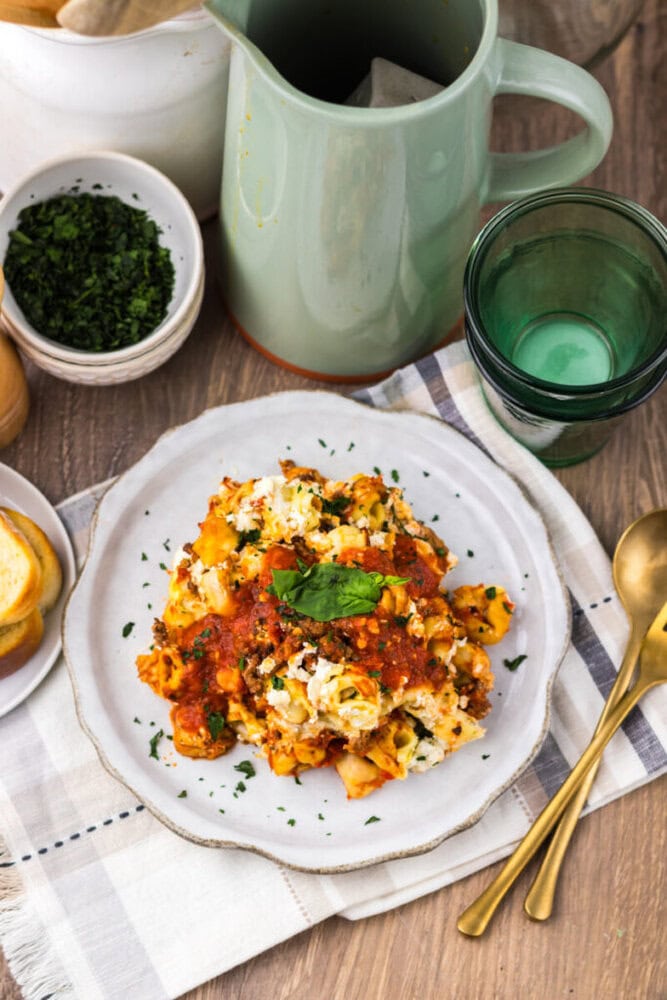 A plate of lasagna garnished with herbs sits on a napkin, next to a bowl of chopped herbs, a green glass, a green pitcher, and gold utensils on a wooden table.