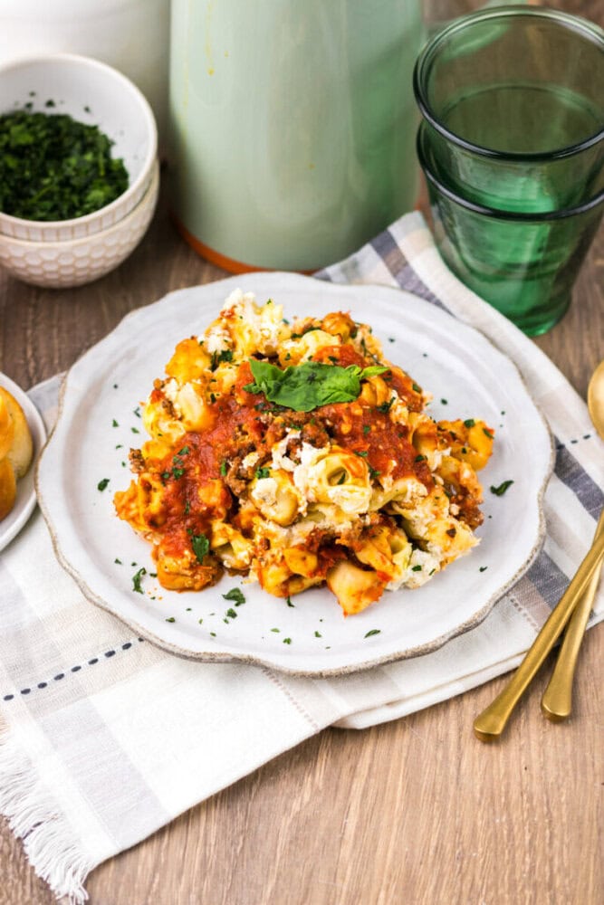 A plate of baked pasta with tomato sauce and cheese, garnished with parsley, sits on a cloth napkin beside gold utensils, glasses, and a bowl of chopped herbs.