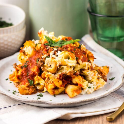 A plate of baked pasta with tomato sauce, ricotta cheese, and basil, served on a white plate with bread, bowls, and green glasses in the background.