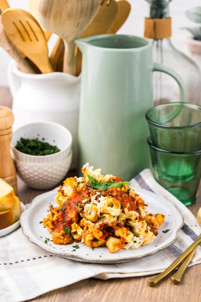 A plate of baked pasta with tomato sauce and cheese sits on a table set with green glasses, bread, and kitchen utensils in the background.