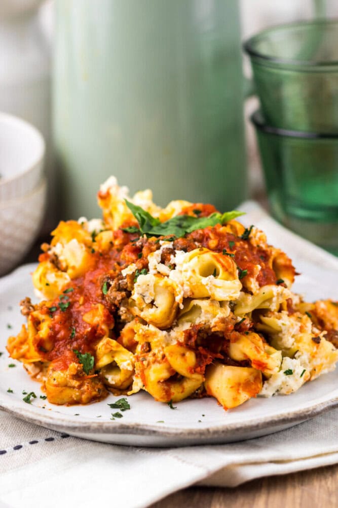A serving of baked pasta with tomato sauce, ground meat, melted cheese, and herbs on a white plate.