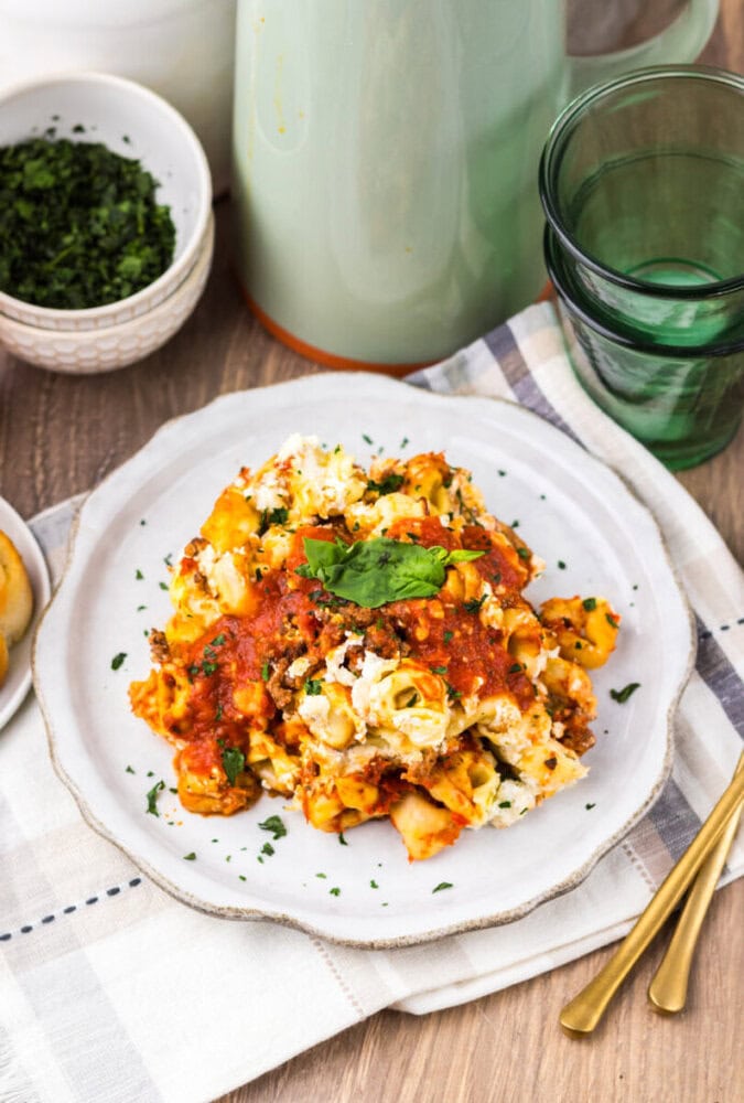 A plate of baked pasta with tomato sauce, cheese, and herbs sits on a cloth napkin next to gold utensils, green glasses, and a bowl of chopped parsley.