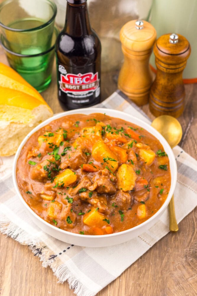 A bowl of beef stew with potatoes and carrots, garnished with parsley, sits on a napkin next to sliced bread, a bottle of root beer, and salt and pepper shakers.