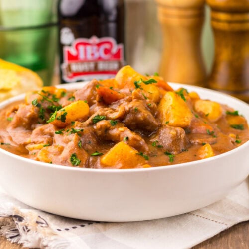 A white bowl filled with beef stew containing chunks of meat, potatoes, and carrots, garnished with herbs, sits on a napkin with bread and a bottle in the background.