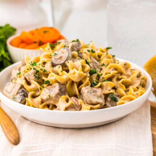 A bowl of creamy beef stroganoff with egg noodles, garnished with parsley, sits on a napkin with bread, carrots, and fresh herbs nearby.