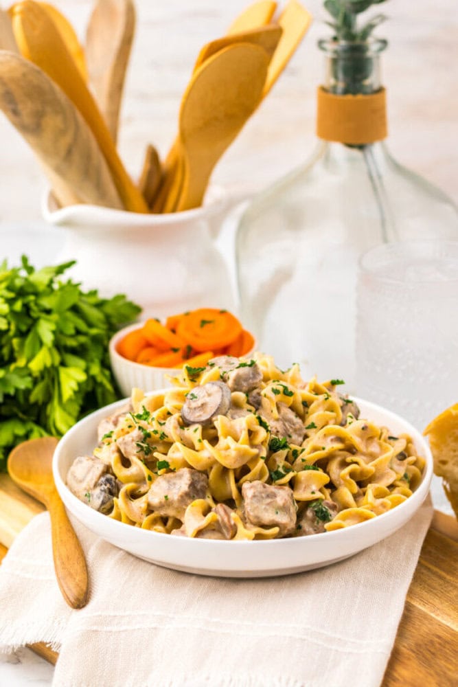 A bowl of creamy beef stroganoff with mushrooms and egg noodles is garnished with parsley. Fresh parsley, sliced carrots, a glass of water, and wooden utensils are in the background.