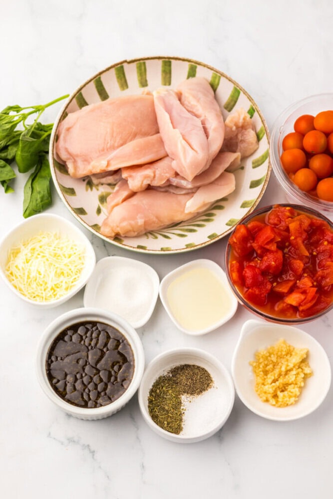 A plate of raw chicken breasts surrounded by bowls of grape tomatoes, canned diced tomatoes, shredded cheese, sugar, oil, chopped garlic, black beans, seasonings, and fresh basil.