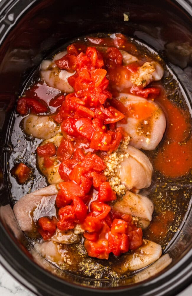 Raw chicken breasts, diced tomatoes, minced garlic, and seasonings in a slow cooker, ready to be cooked.