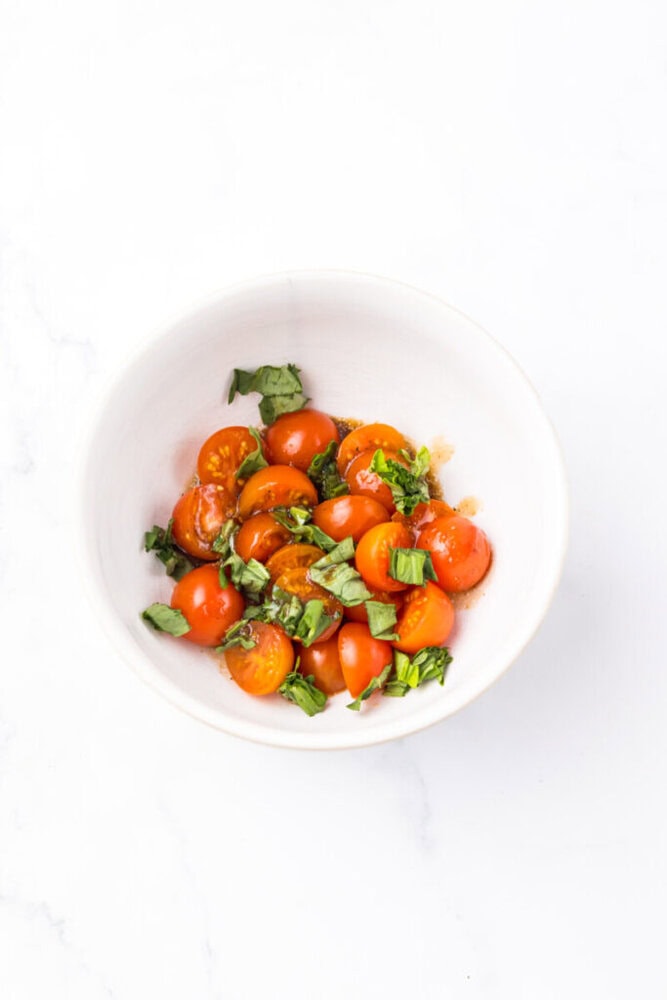 A white bowl filled with halved cherry tomatoes and chopped fresh basil on a white surface.