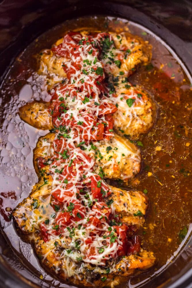 Cooked chicken breasts topped with marinara sauce and melted cheese, garnished with chopped herbs, in a slow cooker with sauce.
