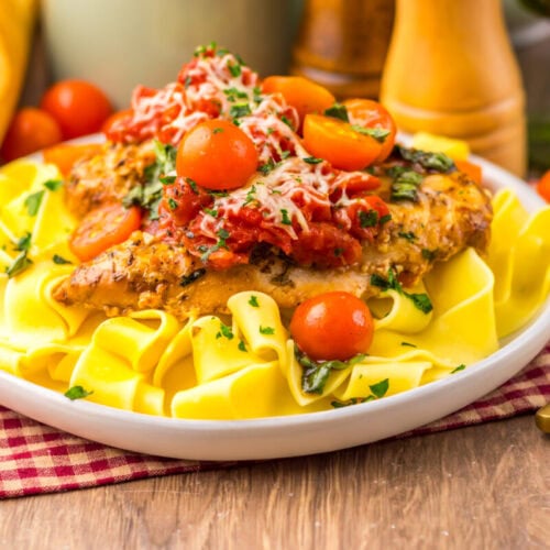 A plate of pappardelle pasta topped with breaded chicken, tomato sauce, cherry tomatoes, grated cheese, and chopped herbs, with bread and utensils in the background.