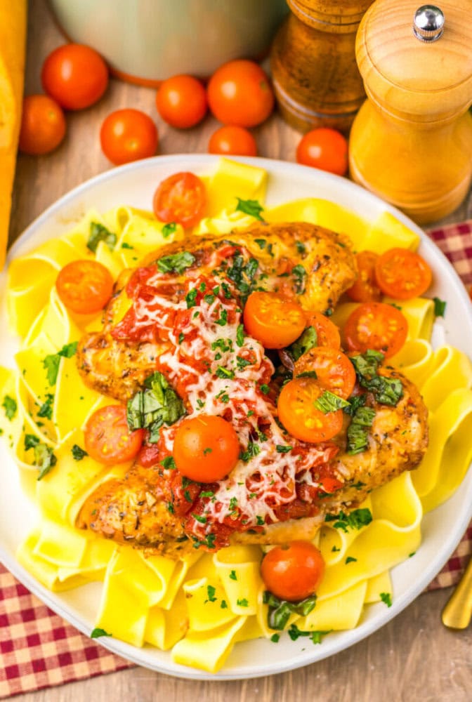 A plate of pappardelle pasta topped with two pieces of seasoned chicken, tomato sauce, cherry tomatoes, shredded cheese, and chopped herbs.
