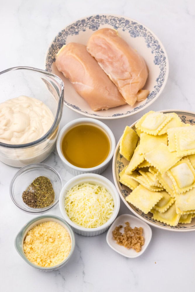 Ingredients on a white surface: raw chicken breasts, ravioli, mayonnaise, grated cheese, parmesan, Italian seasoning, garlic, and olive oil in separate bowls and plates.