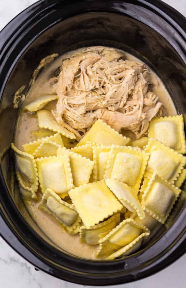 Shredded chicken, sauce, and uncooked ravioli in a slow cooker, ready to be cooked together.