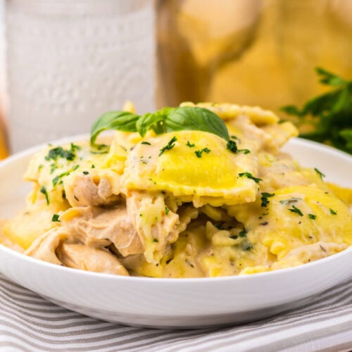 A white bowl of ravioli pasta in a creamy sauce, garnished with chopped herbs and a basil leaf, sits on a striped napkin next to bread and fresh parsley.