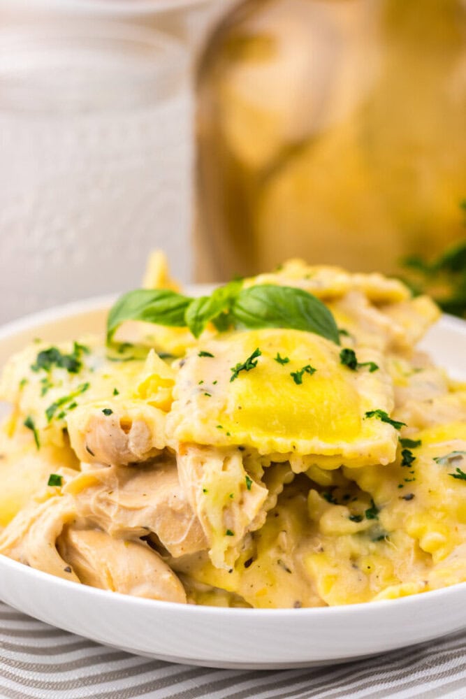 A bowl of ravioli pasta in a creamy sauce with pieces of chicken, garnished with chopped parsley and a sprig of basil.