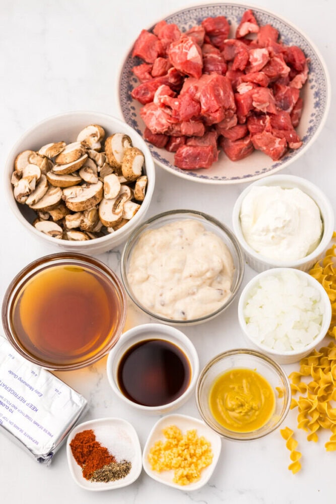 Overhead view of ingredients for beef stroganoff, including cubed beef, sliced mushrooms, broth, cream, onions, egg noodles, seasonings, garlic, mustard, Worcestershire sauce, and soup.