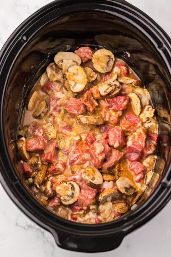 A slow cooker filled with a mixture of raw beef chunks, sliced mushrooms, and diced tomatoes in a creamy sauce.