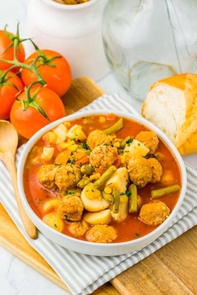 Bowl of vegetable stew with meatballs, potatoes, and green beans, next to tomatoes and bread on a striped cloth.