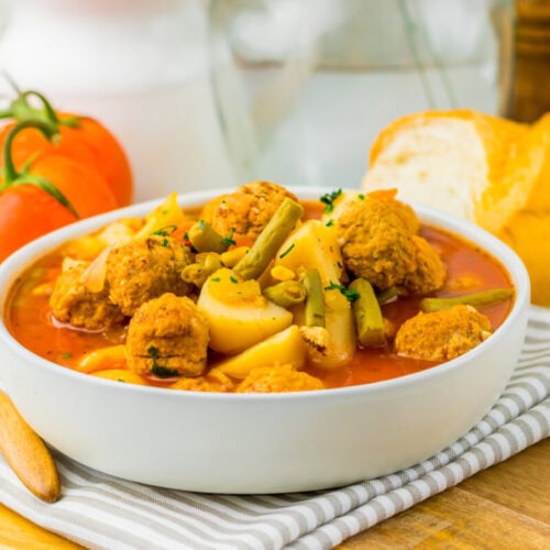 Bowl of meatball stew with potatoes and green beans, bread, tomatoes, and a wooden spoon on the side.