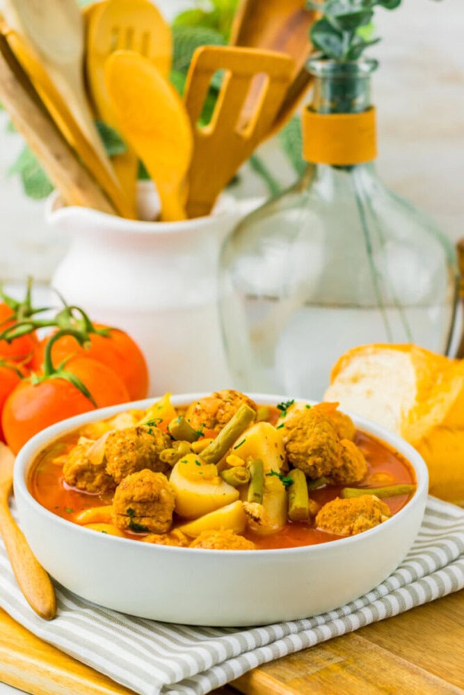 A bowl of vegetable stew with potatoes, green beans, and bread, set on a striped napkin with tomatoes in the background.
