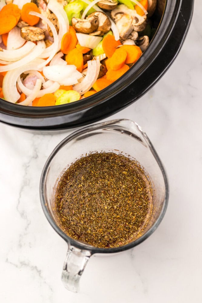 A glass measuring cup with herb seasoning mix next to a slow cooker filled with sliced onions, carrots, celery, and mushrooms.