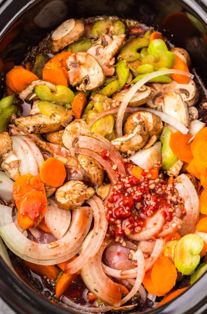 Sliced mushrooms, carrots, celery, onions, and minced garlic on top of raw chicken in a slow cooker with sauce.