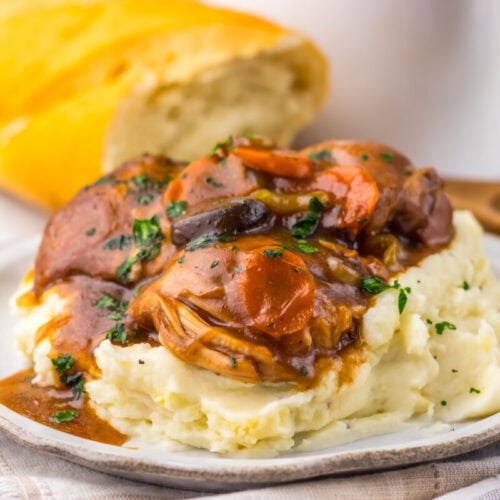 A plate of mashed potatoes topped with pot roast, carrots, and gravy, with a loaf of bread in the background.