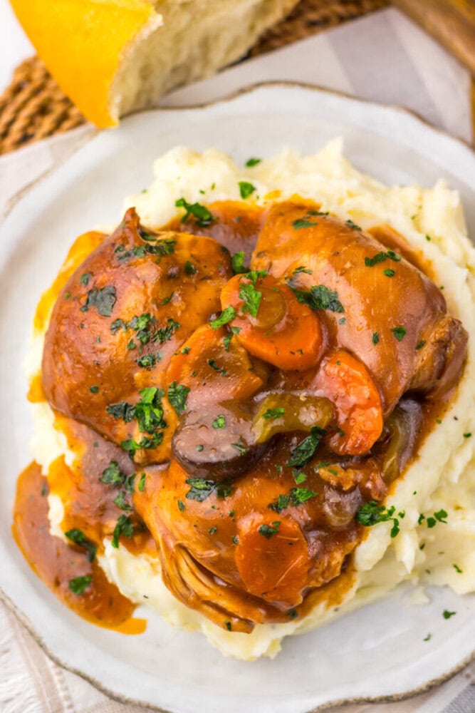 Braised chicken thighs with carrots, mushrooms, and sauce served over mashed potatoes on a white plate, garnished with chopped herbs.