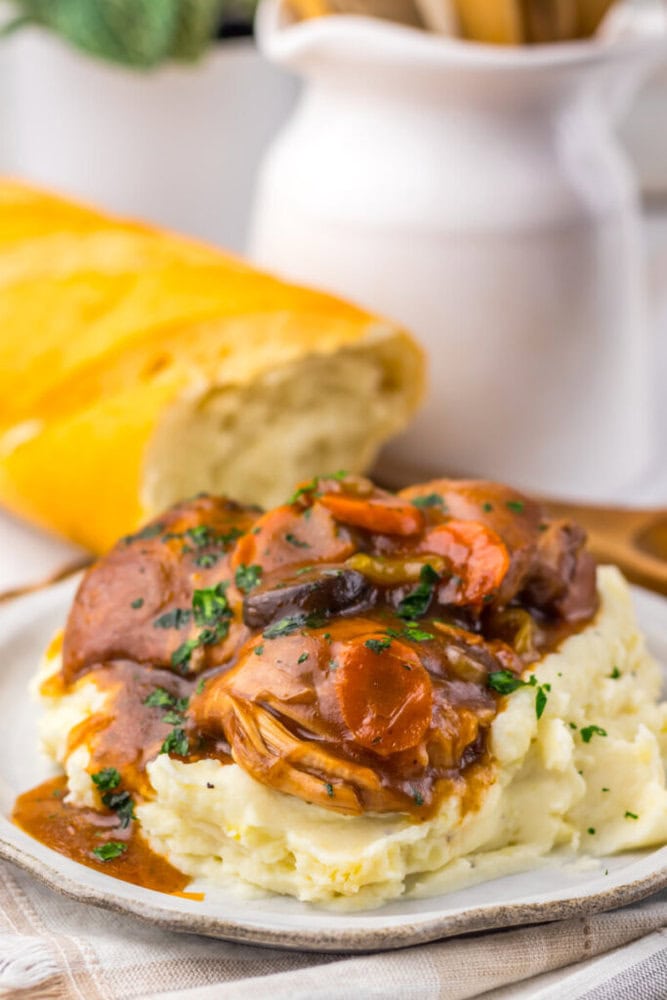 A plate of mashed potatoes topped with beef stew and vegetables, garnished with herbs, with a loaf of bread in the background.