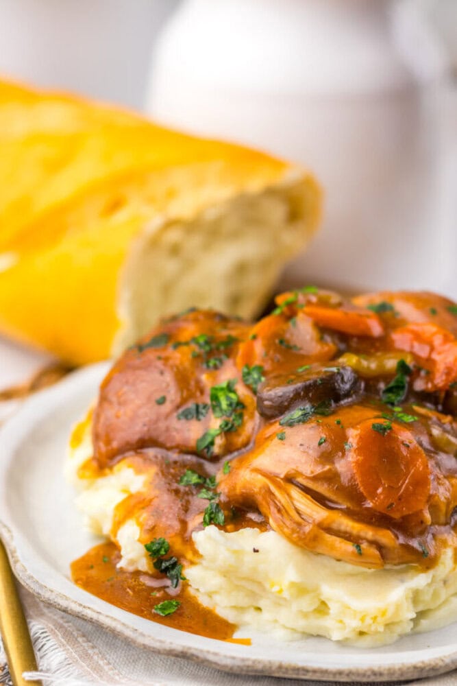 Plate of mashed potatoes topped with sliced sausage, vegetables, and brown gravy, with a loaf of bread in the background.