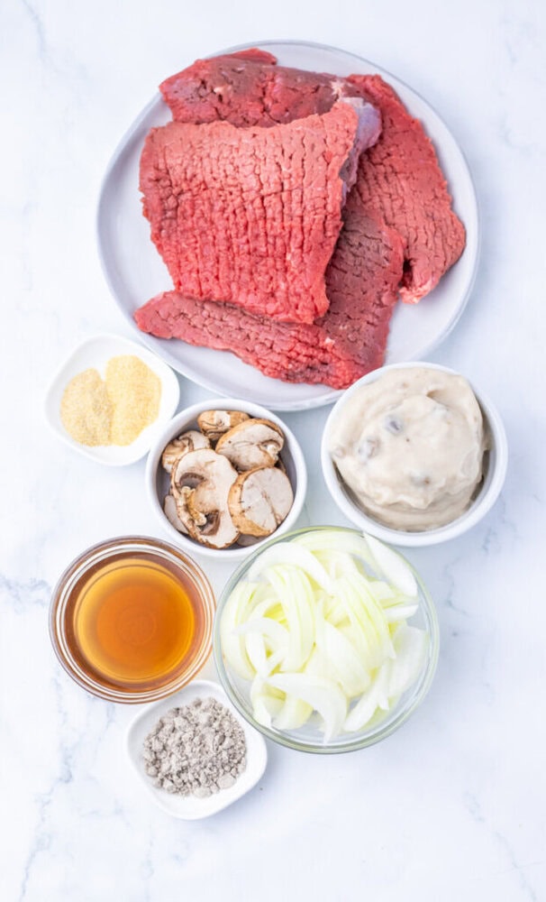 Top-down view of raw cube steak on a plate with separate bowls of sliced onions, sliced mushrooms, cream of mushroom soup, beef broth, garlic powder, and pepper on a white surface.