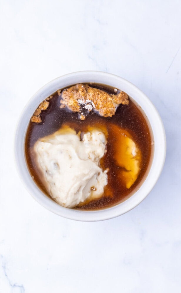 A bowl containing brown liquid, a scoop of white dough-like substance, and visible granules on top, all on a white background.