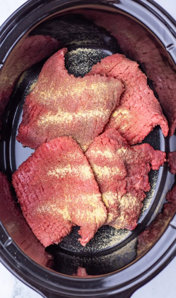 Raw pieces of seasoned beef placed inside a black slow cooker, seen from above.