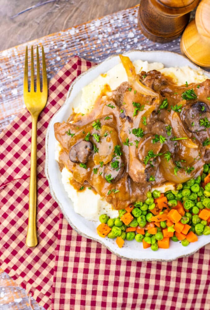 Plate of pork chops with onions and gravy, mashed potatoes, peas, carrots, and a gold fork on a checkered napkin.