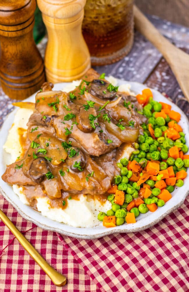 A plate of mashed potatoes topped with sliced beef and mushroom gravy, served with a side of mixed peas and diced carrots. Salt and pepper shakers are in the background.