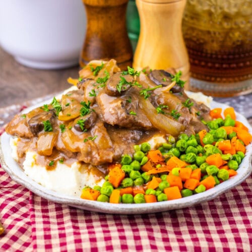 A plate of beef with brown gravy and onions served over mashed potatoes, accompanied by mixed peas and diced carrots on a red checkered cloth.