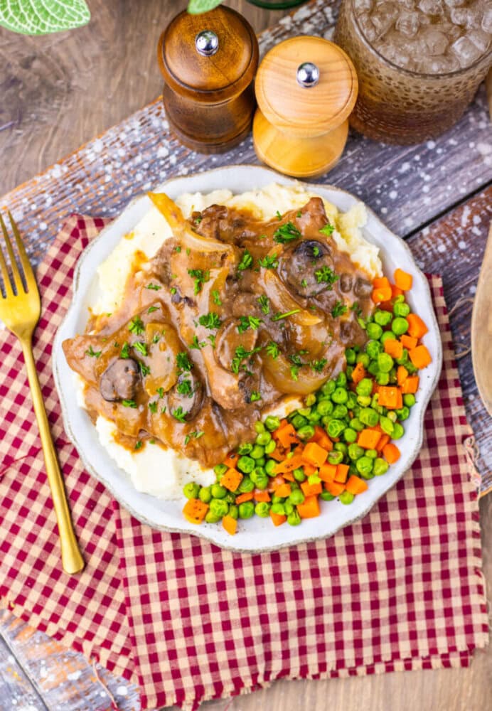 Plate of mashed potatoes topped with beef, onions, and gravy, served with mixed peas and carrots on a red checkered napkin, with a gold fork and drink nearby.